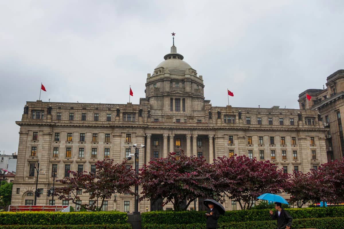 Grand bâtiment néoclassique avec colonnes et dôme, arborant des drapeaux chinois, vu depuis une rue animée et un espace paysager par temps nuageux