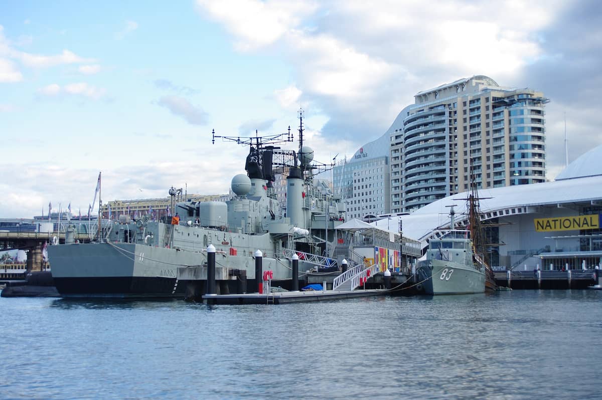 Widok z zewnątrz na Australijskie Narodowe Muzeum Morskie z historycznymi statkami i łodziami zacumowanymi w Darling Harbour w Sydney, pod błękitnym niebem.