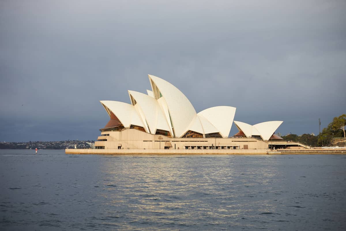 Sydney Opera House viewed from across the harbor, its iconic white sails illuminated against a cloudy sky, with the building reflected in calm water.