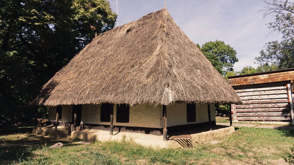 Historisches Bauernhaus mit Strohdach, umgeben von Grünpflanzen und Bäumen in einem Freilichtmuseum, unter dem Tageshimmel.
