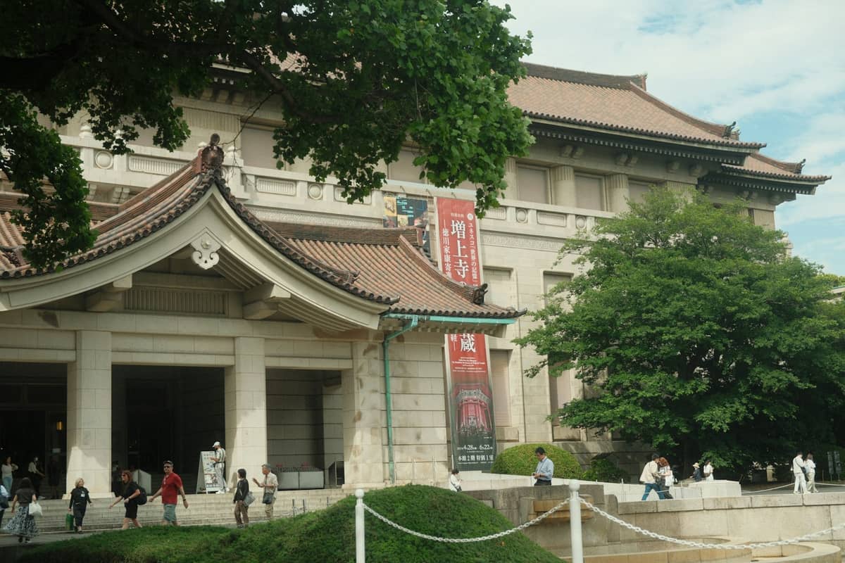 Exterior of the Tokyo National Museum Honkan building with visitors walking outside, traditional Japanese roof elements, and banners at the entrance.