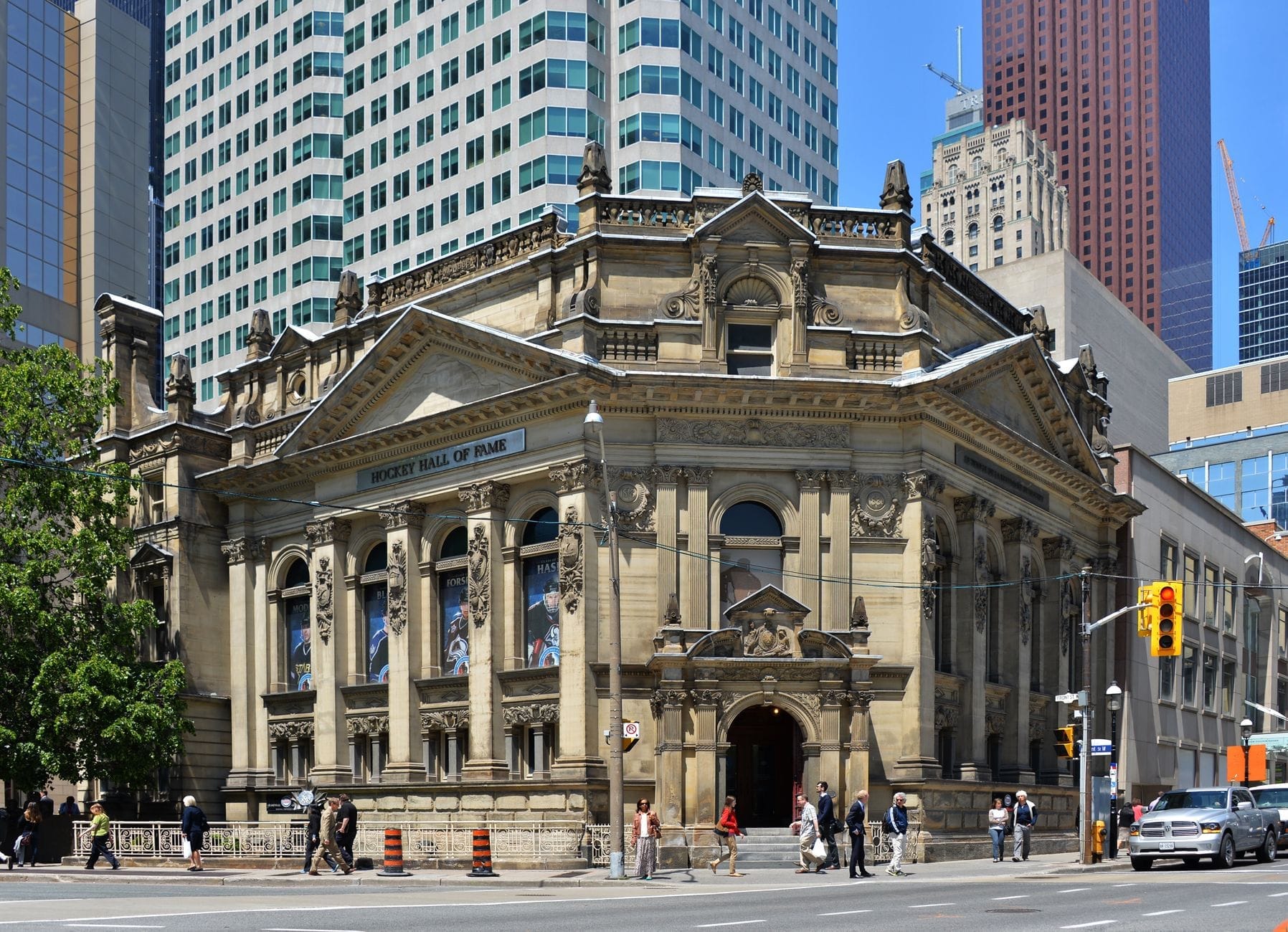 Histórico edifício de pedra numa movimentada esquina de Toronto, com faixas do Hockey Hall of Fame, pedestres do lado de fora e arranha-céus modernos ao fundo.