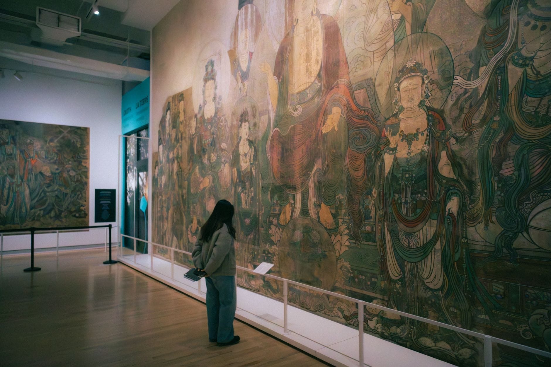 Um visitante admira um grande mural detalhado com figuras asiáticas tradicionais em uma galeria bem iluminada de um museu em Toronto.