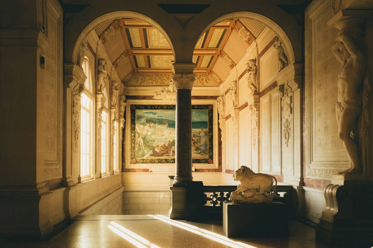 Eleganter, sonnendurchfluteter Galeriesaal mit Bogenfenstern, klassischen Säulen, Marmor-Löwenstatue und venezianischem Renaissance-Gemälde an der Wand.