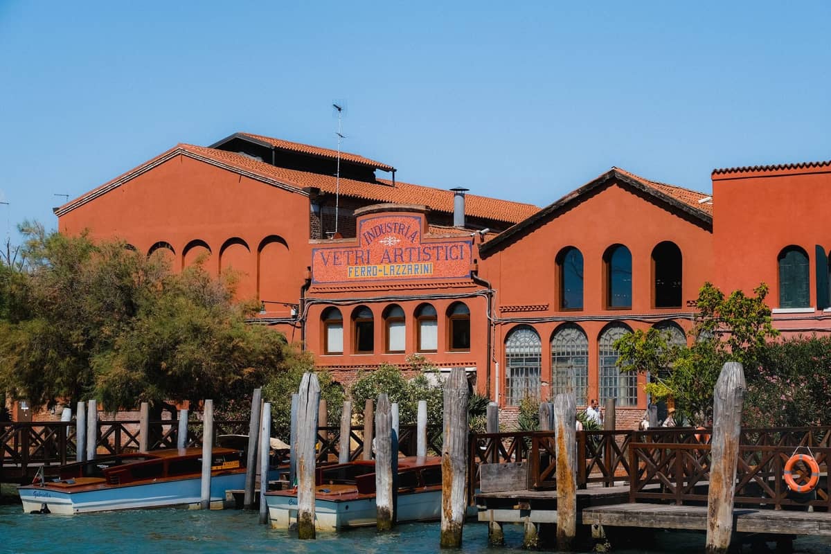 Historisches Glasfabrikgebäude von Murano am Ufer, mit rot-oranger Fassade, Bogenfenstern und Holzdocks am Kanal.