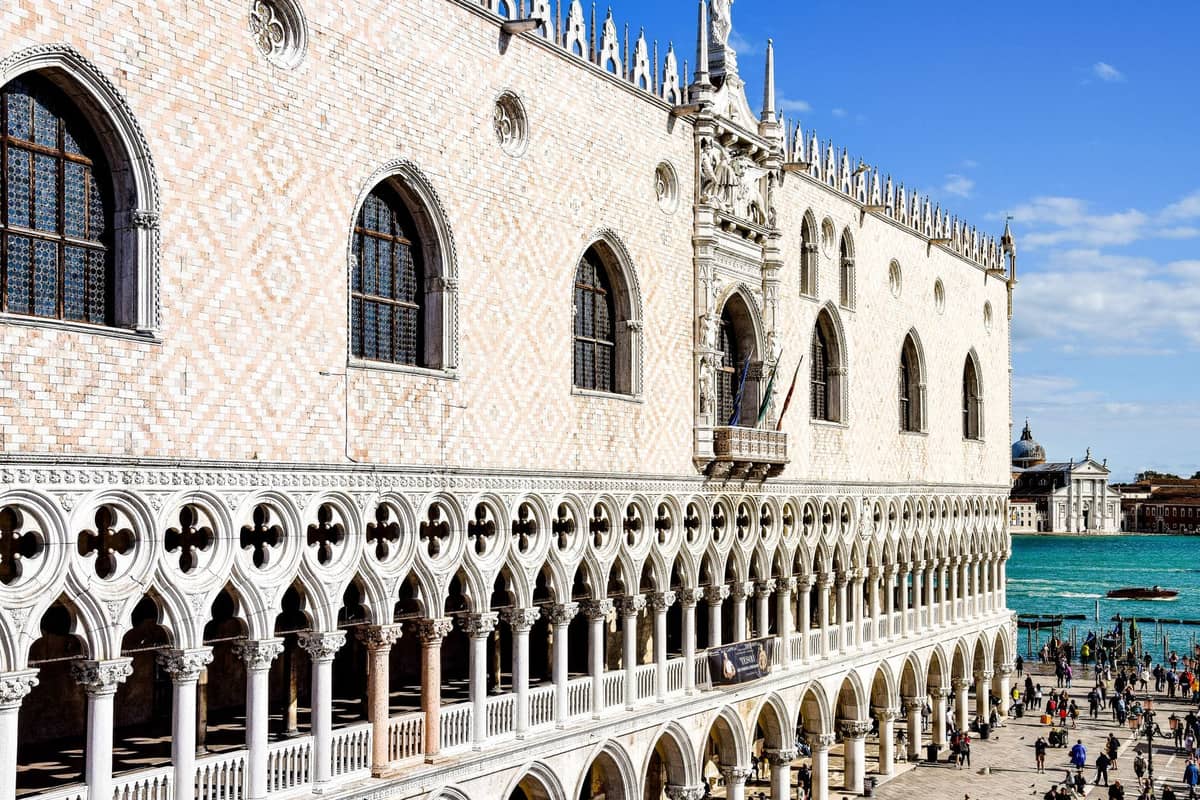 Weitblick auf den Dogenpalast in Venedig mit seinen charakteristischen gotischen Bögen, der aufwendigen Fassade und Menschen, die im Vordergrund nahe dem Canal Grande spazieren.