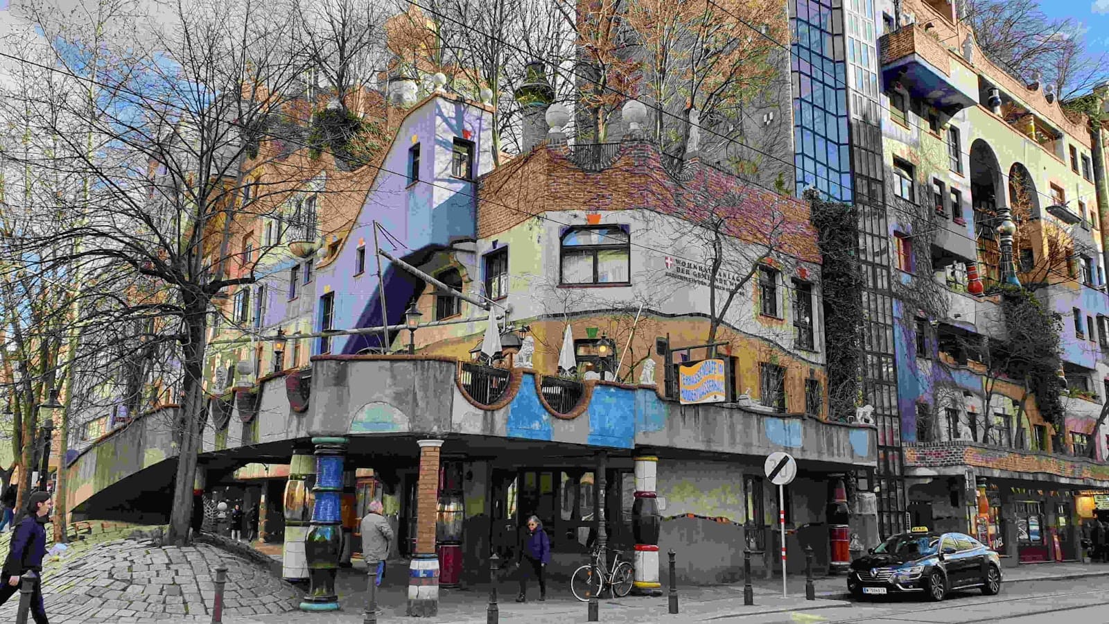Widok narożny kolorowego Hundertwasserhaus w Wiedniu z pieszymi, drzewami i ruchem ulicznym przed budynkiem.