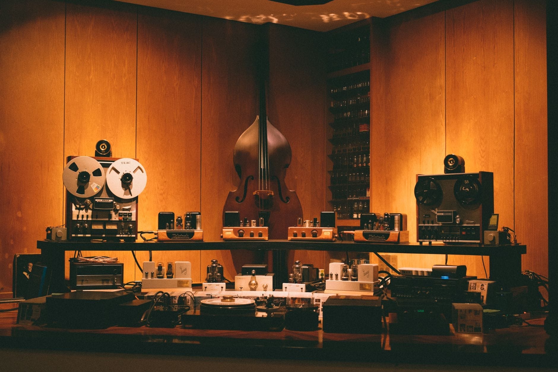 Sala de estudio de música con luz cálida, máquinas de cinta de carrete a carrete, contrabajo y equipos de audio vintage expuestos contra una pared de paneles de madera.