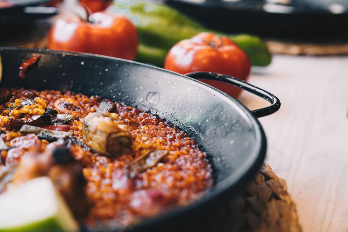 Primo piano di una paella tradizionale in una padella, con riso, pezzi di carne e verdure, con pomodori sullo sfondo.