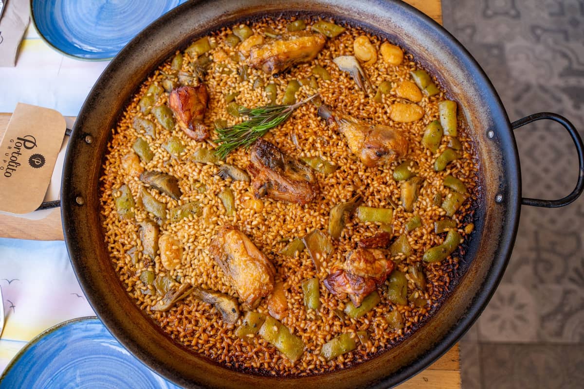 Una tradizionale paella valenciana in una padella rotonda e larga, con riso, pollo, coniglio, fagioli e rosmarino su un tavolo apparecchiato a Valencia.
