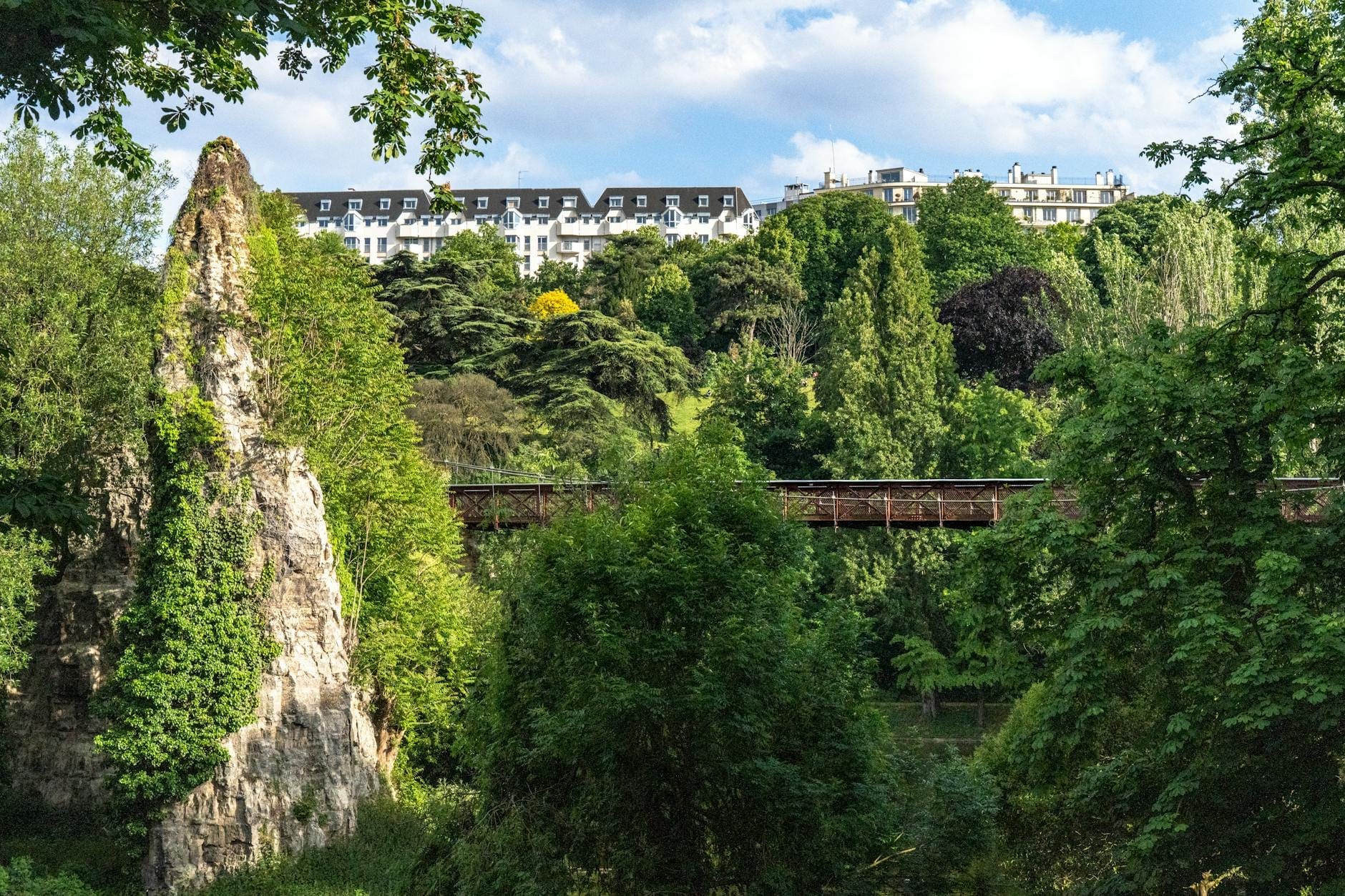 Dramatischer grüner Park in Paris mit steilem Felsvorsprung, Fußgängerbrücke, üppigen Bäumen und Pariser Wohngebäuden im Hintergrund.