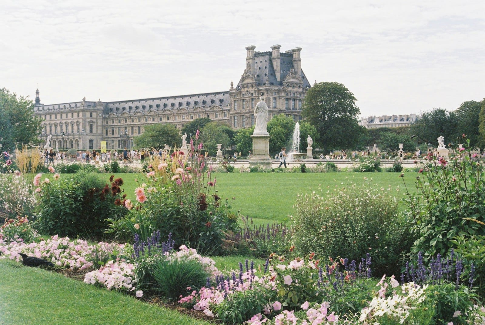 Rigoglioso giardino parigino con aiuole colorate e statue, sullo sfondo la classica architettura francese sotto un cielo parzialmente nuvoloso.