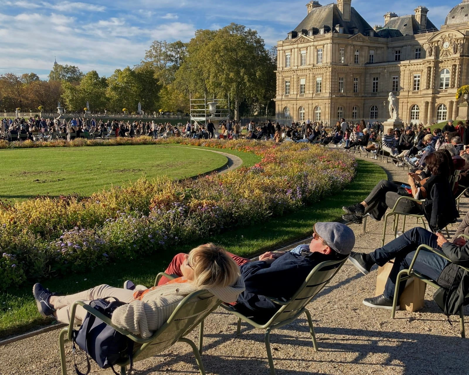 Menschen, die sich auf grünen Stühlen um einen gepflegten Garten in der Nähe eines eleganten historischen Gebäudes in einem Pariser Park unter blauem Himmel entspannen.