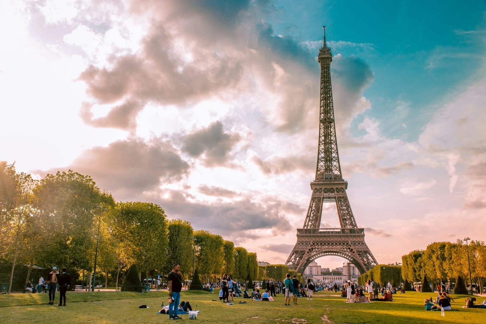Menschen beim Picknick auf dem grünen Rasen des Champ de Mars mit dem Eiffelturm im Hintergrund an einem hell bewölkten Tag