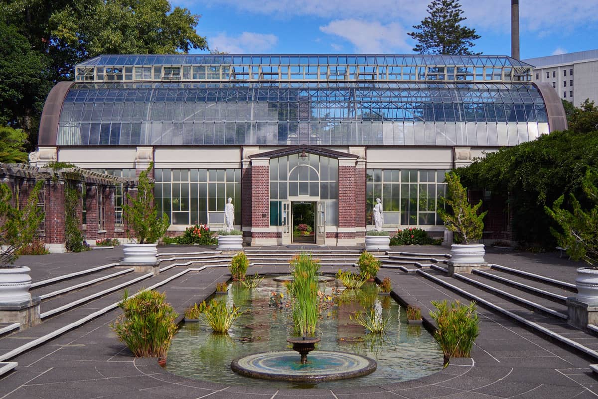 Extérieur des Wintergardens de l'Auckland Domain avec serre, bassin formel, statues et verdure luxuriante sous un ciel bleu.