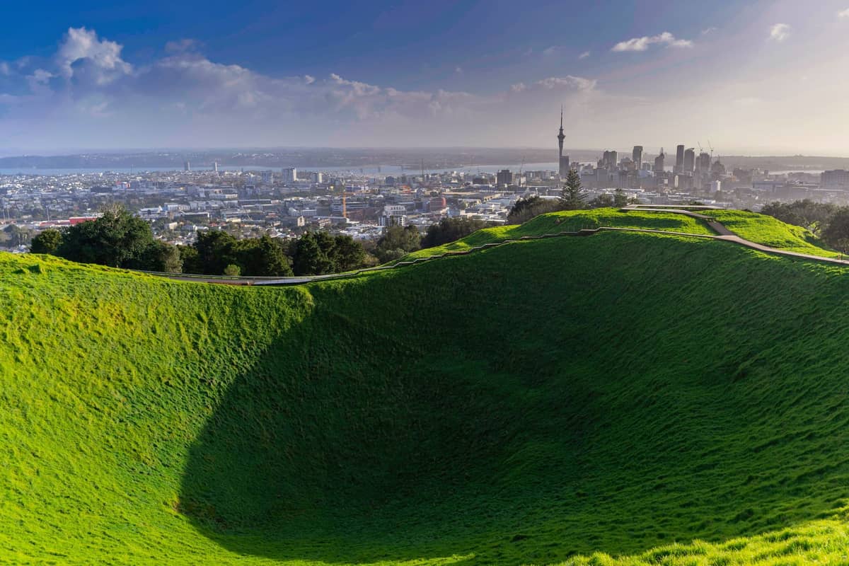 Blick auf den Vulkankrater des Mount Eden mit sattem grünen Gras und der Skyline von Auckland im Hintergrund unter blauem Himmel.