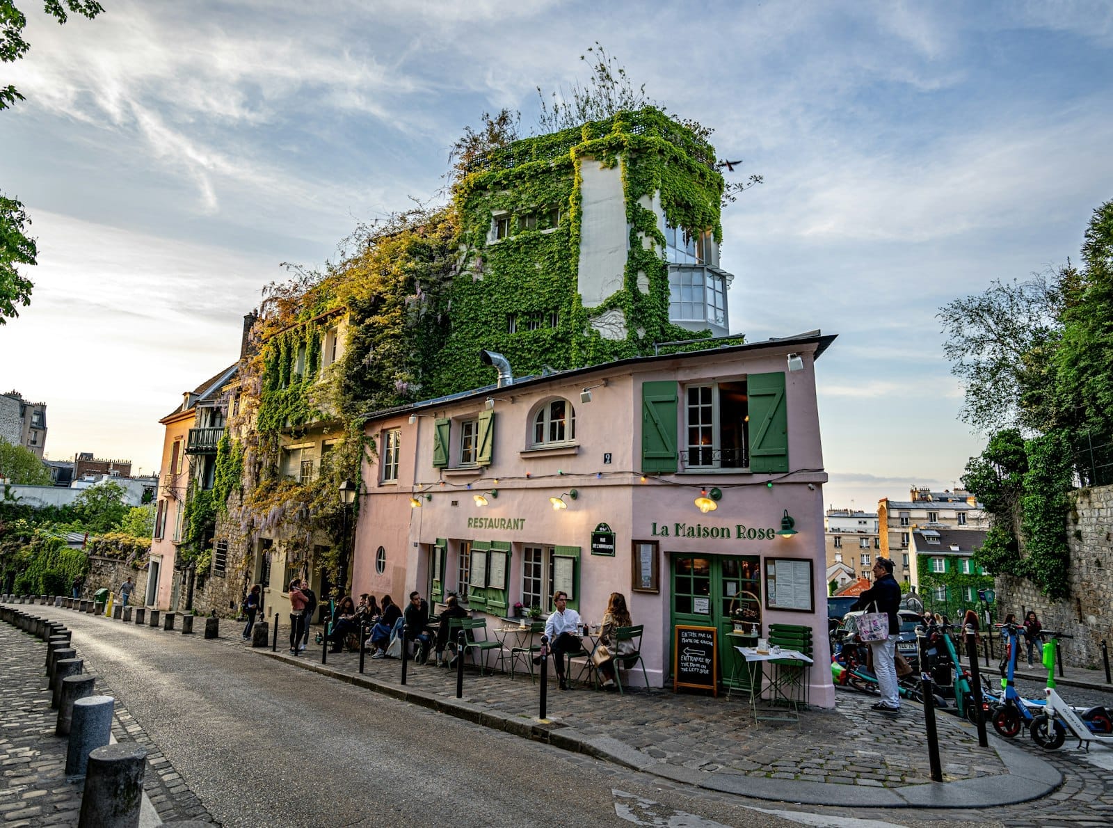 Charmantes, efeubewachsenes Café La Maison Rose in einer ruhigen Kopfsteinpflasterstraße in Montmartre, mit speisenden und schlendernden Menschen bei Sonnenuntergang.