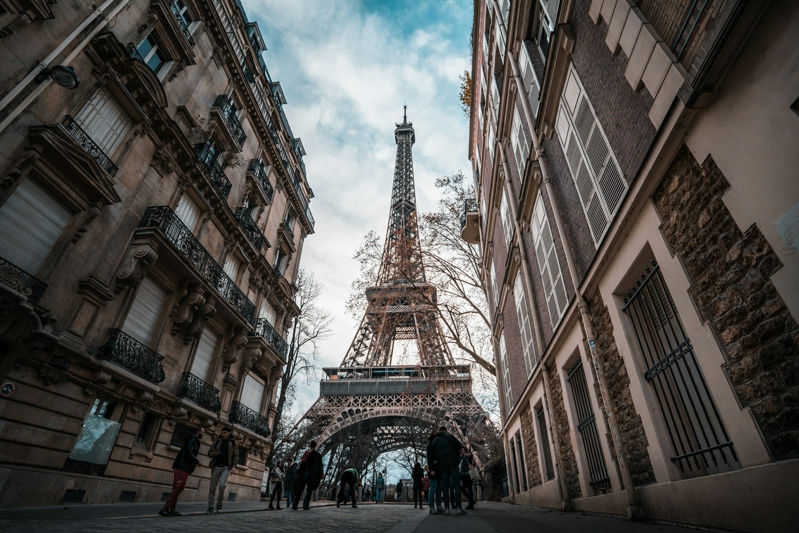 Vista della Torre Eiffel incorniciata dai classici palazzi parigini, con persone che camminano lungo la strada in una giornata nuvolosa.