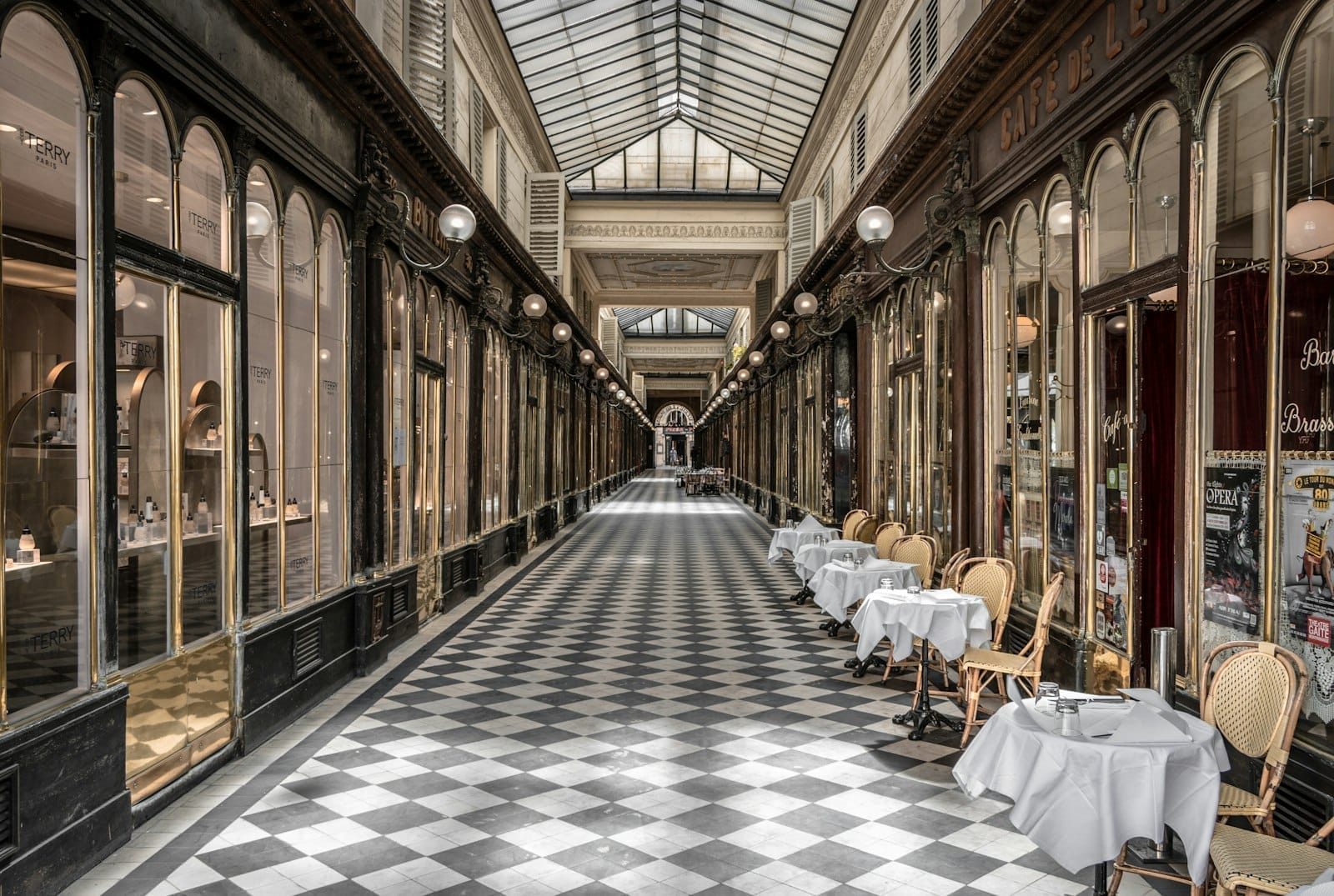 Überdachte Passage in Paris mit Glasdach, Mosaikboden, eleganten Geschäften und aufgestellten Tischen auf einer Seite, typisch für die Arkaden des 19. Jahrhunderts.