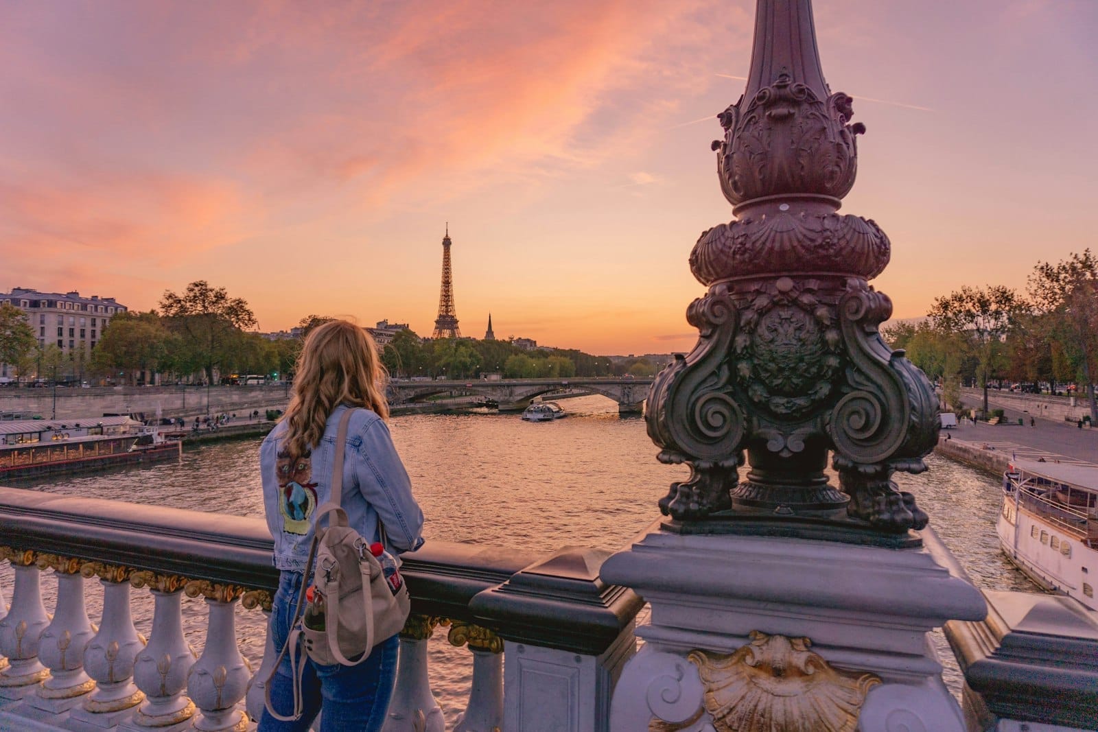 Frau auf einer verzierten Brücke in Paris bei Sonnenuntergang mit Blick auf die Seine, dem Eiffelturm im Hintergrund und dekorativen Laternen.
