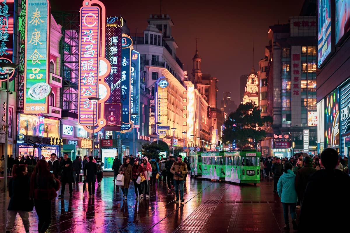 Nanjing Road en Shanghái de noche con multitudes de personas caminando entre tiendas iluminadas con neón y luces de colores reflejadas en el pavimento mojado.
