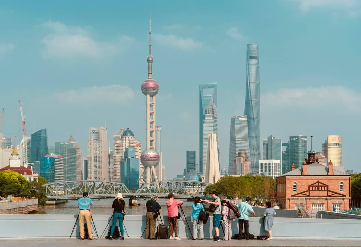 Menschen mit Kameras stehen auf einer Brücke mit Blick auf Shanghais Skyline – mit dem Bund, dem Suzhou Creek und den Wolkenkratzern von Lujiazui unter einem klaren blauen Himmel.