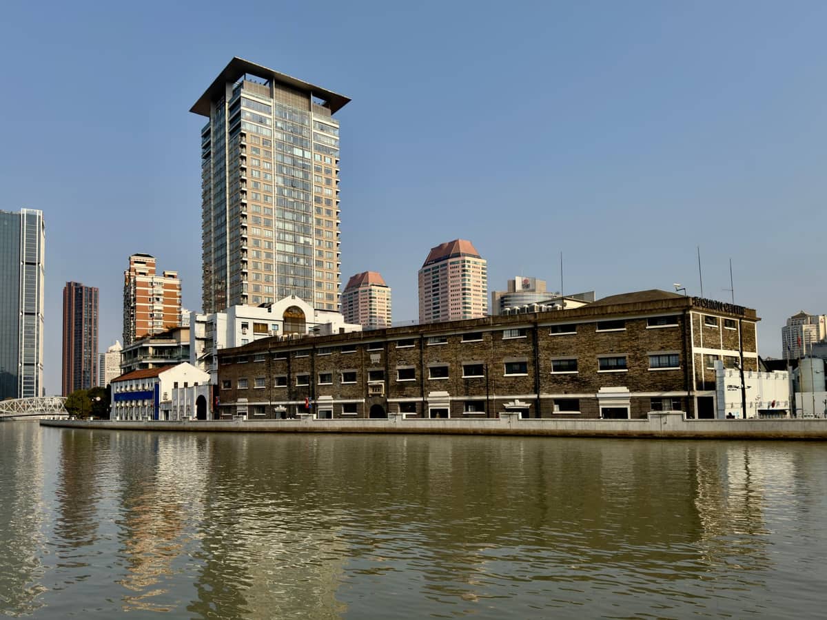 Vista del Suzhou Creek en Shanghái con antiguos edificios de almacenes industriales reconvertidos y modernos rascacielos a lo largo del paseo marítimo bajo un cielo despejado.