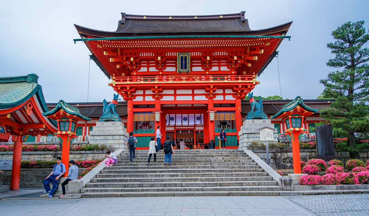 People walking up the stone steps toward the vibrant red Romon gate of Fushimi Inari Taisha, a famous Shinto shrine in Kyoto.