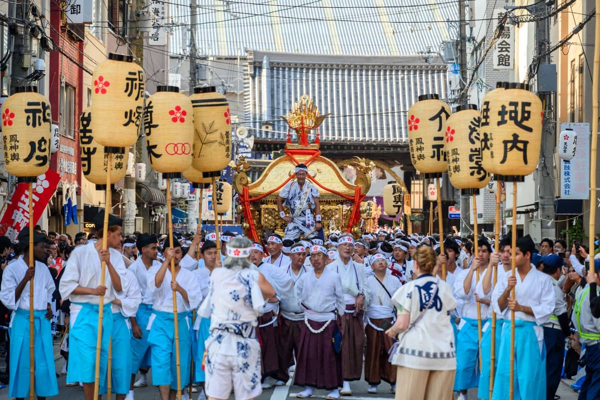 Japanese festival procession in Osaka with participants carrying lanterns and a mikoshi through crowded streets near a shrine