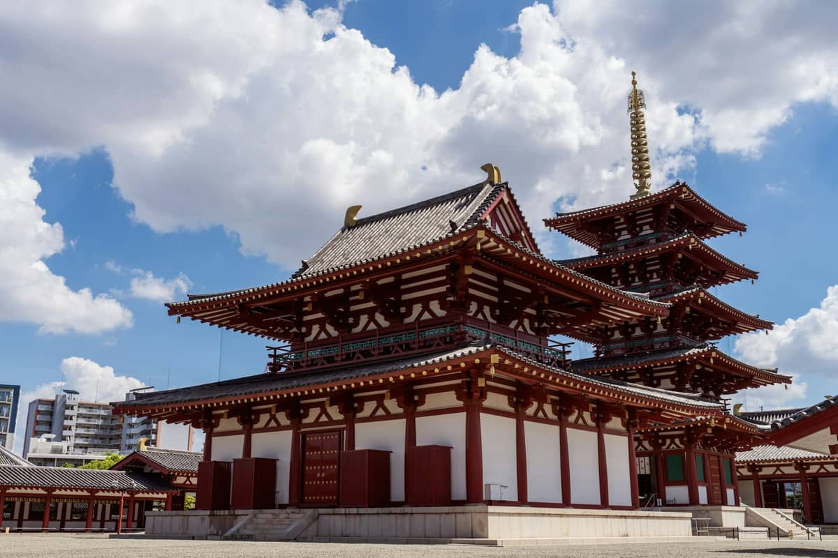 Landscape view of Shitennoji Temple in Osaka, Japan with its distinctive red and white pagoda and surrounding temple buildings under a blue sky with clouds.