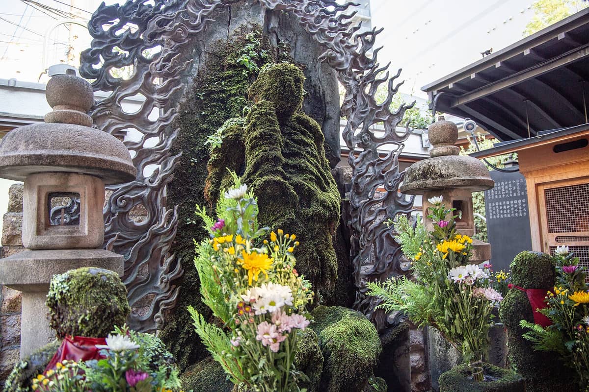 Moss-covered Fudo Myo-o statue at Hozenji Temple in Osaka, surrounded by stone lanterns and fresh flower offerings, in a small shrine setting.