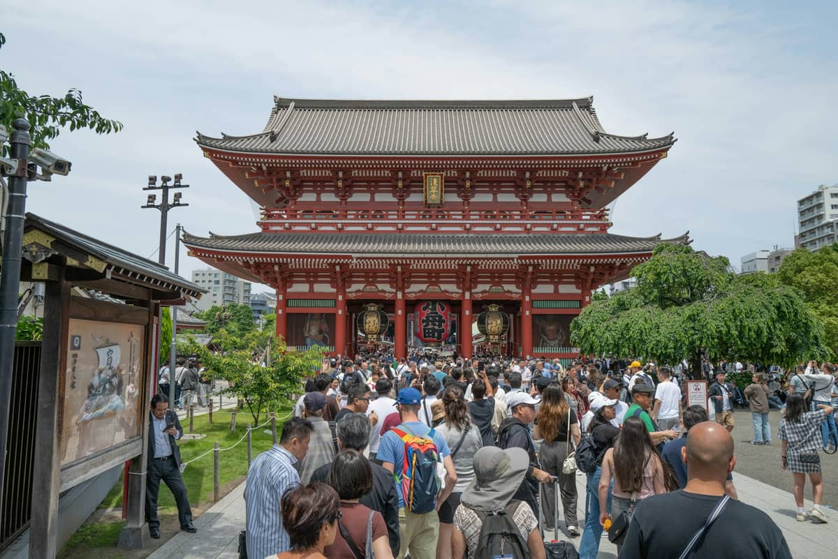 Large crowd approaches the iconic red Hozomon gate of Senso-ji temple in Tokyo on a bright day, capturing a classic temple experience.