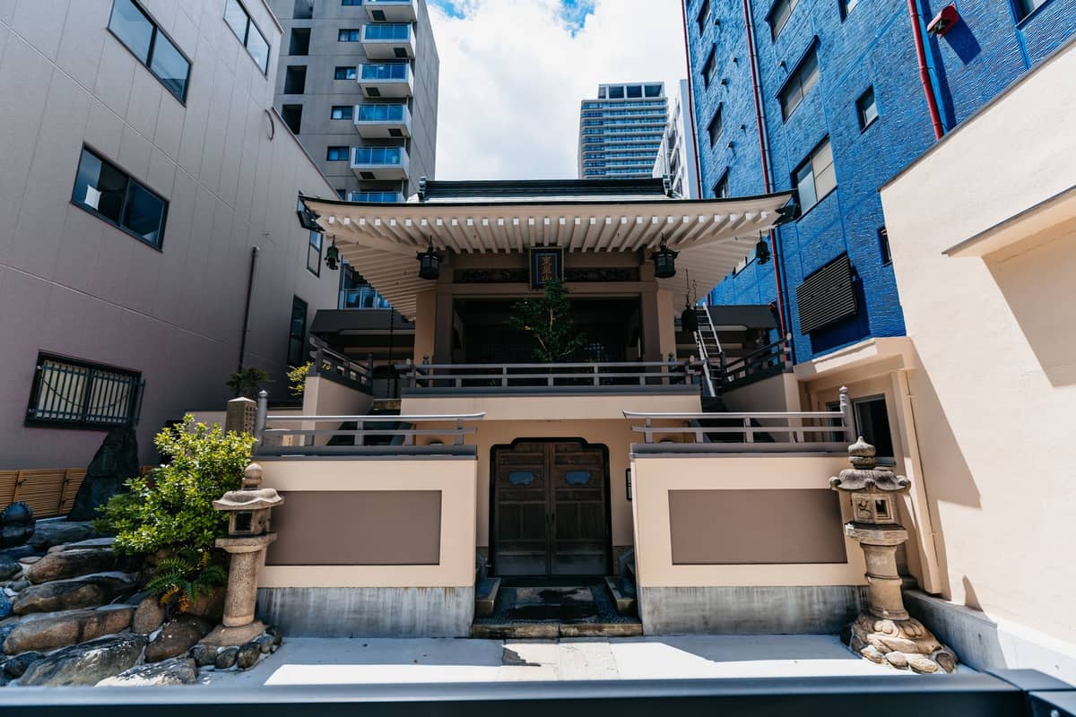 Small neighborhood temple nestled between modern buildings in Tokyo with stone lanterns and greenery at the entrance.