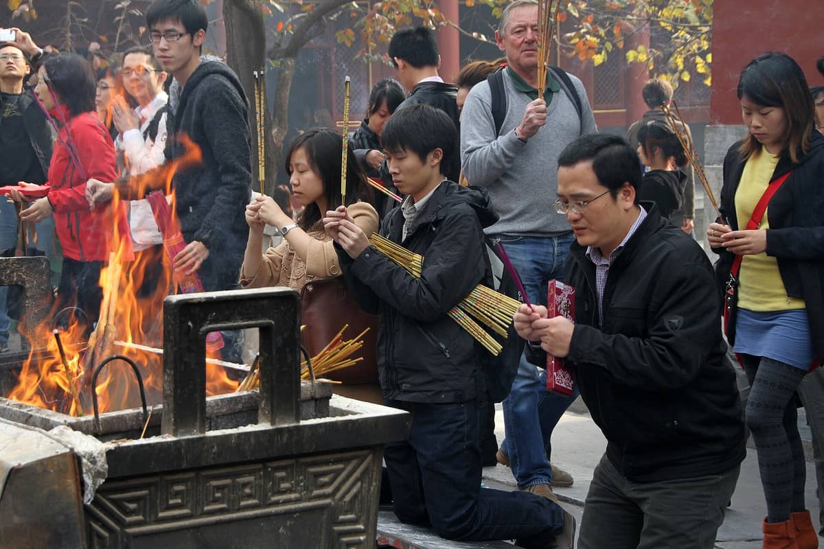 Multitud de fieles con varillas de incienso rezando y haciendo ofrendas en el concurrido patio de un templo budista de Beijing con fuego y humo visibles.