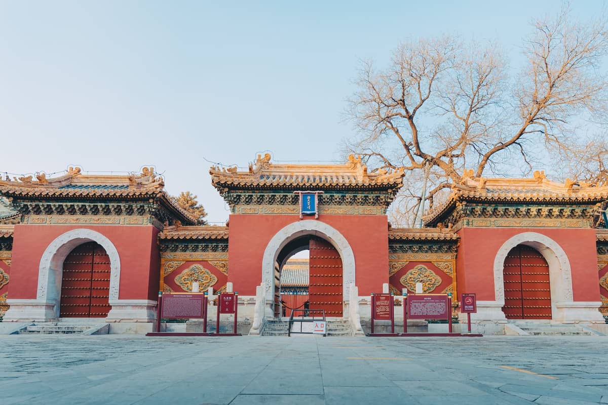 Vista simétrica de las puertas de entrada a un complejo de templos de Beijing con muros rojos, techos ornamentados y árboles sin hojas al fondo.