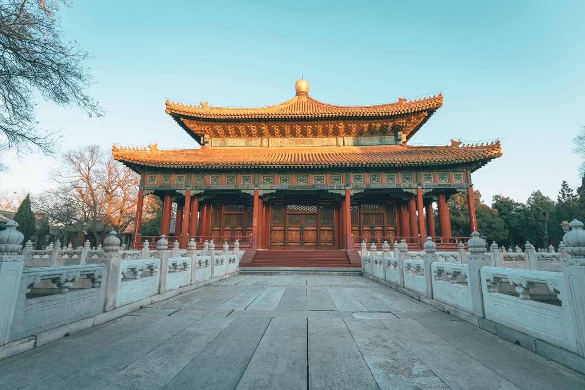 Symmetrischer Blick auf eine traditionelle chinesische Tempelhalle in Peking mit gelbem Ziegeldach, roten Säulen und verziertem Eingangstor, erreichbar über weiße Steinbrücken.