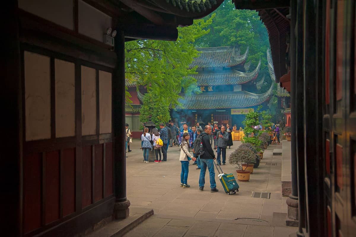 Innenhof eines traditionellen chinesischen buddhistischen Tempels in Chengdu mit Besuchern und aufsteigendem Weihrauchrauch nahe verzierten Dächern.