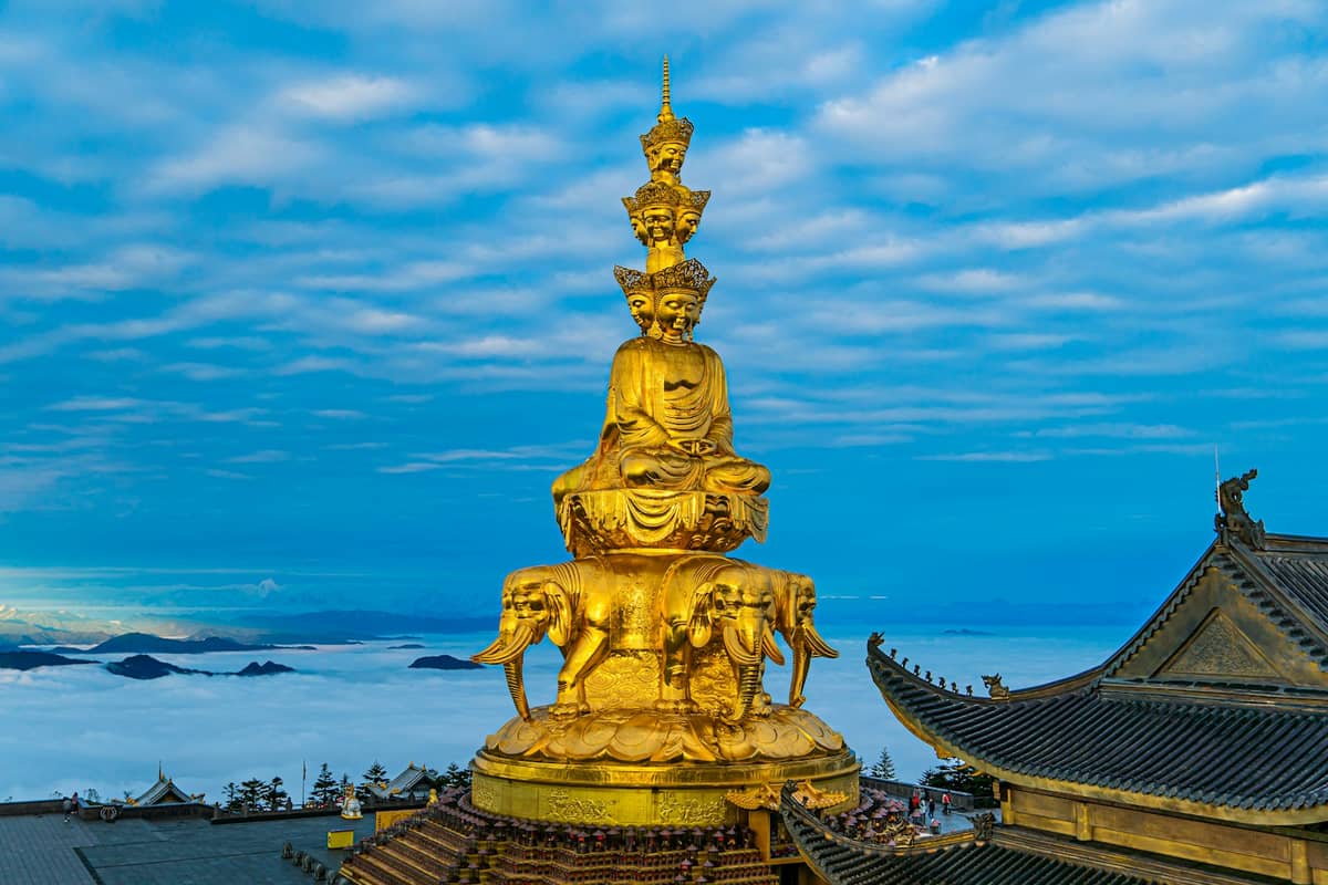 Goldene vielgesichtige Bodhisattva-Statue auf dem Gipfel des Mount Emei mit Tempeldächern und Wolken darunter, unter einem blauen Himmel in Sichuan, China.