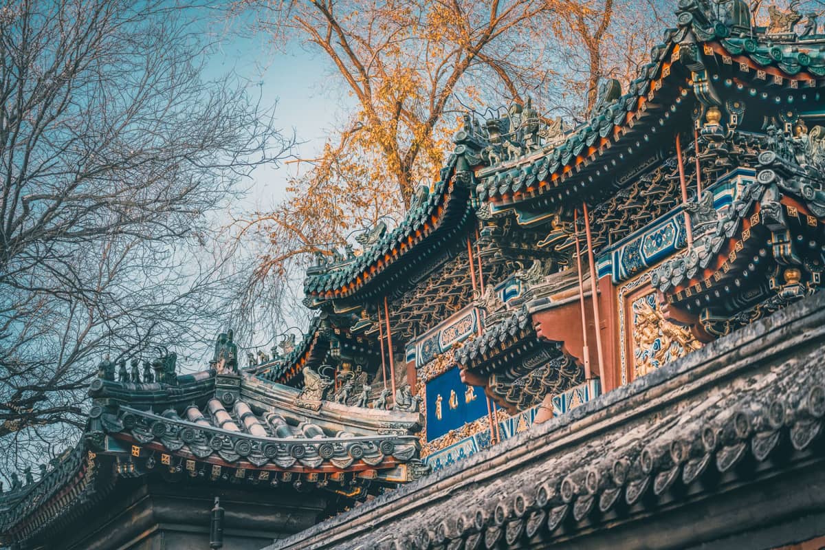 Aufwendig verzierte Dachlinien eines traditionellen chinesischen Tempels, mit farbenfrohen Details und kahlen Bäumen im Hintergrund.