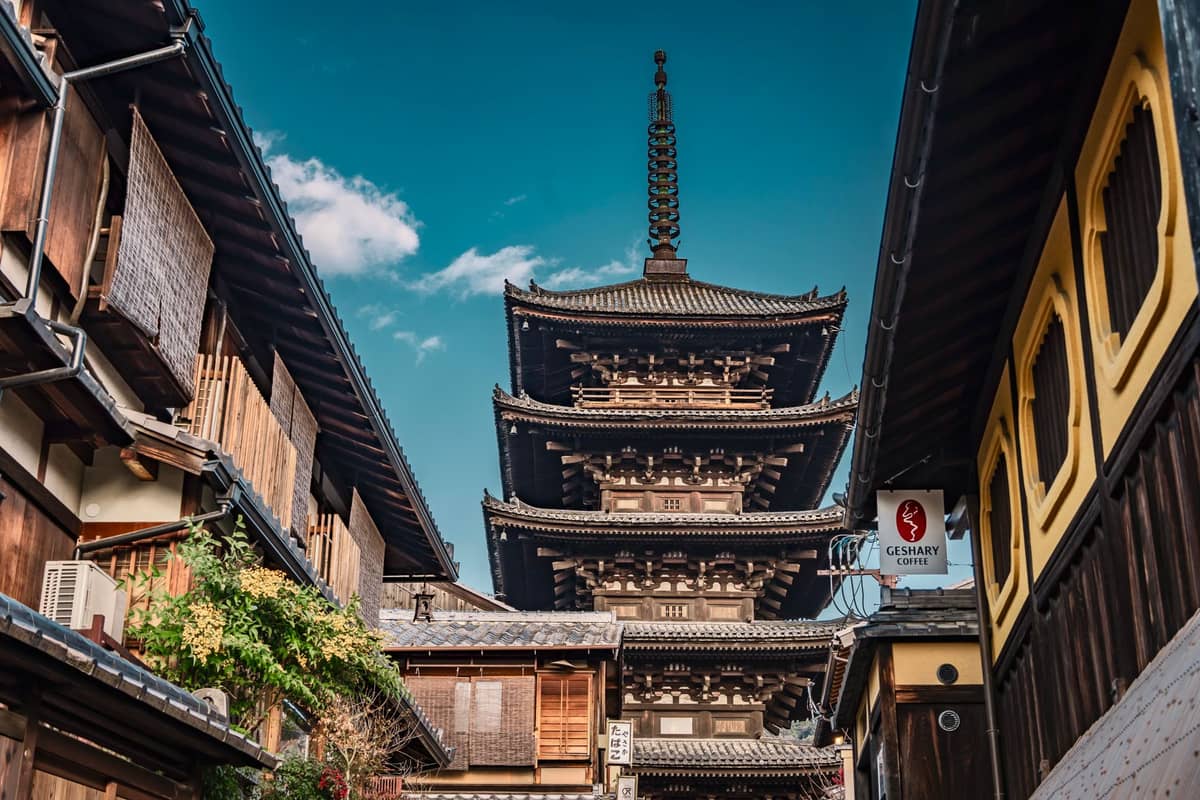 A dramatic view along a Kyoto street framing the ornate five-story Yasaka Pagoda against a blue sky, surrounded by traditional wooden buildings.