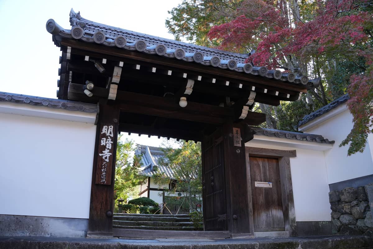 Traditional wooden temple gate with Japanese characters, white walls, and autumn foliage in a quiet, secluded area in Kyoto.