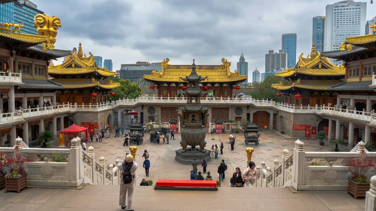 Weitwinkelperspektive auf die goldenen Dächer des Jing'an-Tempels, den großen Innenhof und den Räucherofen, mit Besuchern auf dem Gelände und modernen Shanghaier Wolkenkratzern im Hintergrund.