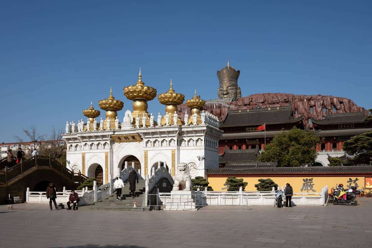 Vista ampia del complesso del tempio buddista di Shanghai con cancello bianco e dorato, mura gialle e grande statua di Buddha sullo sfondo sotto un cielo sereno.