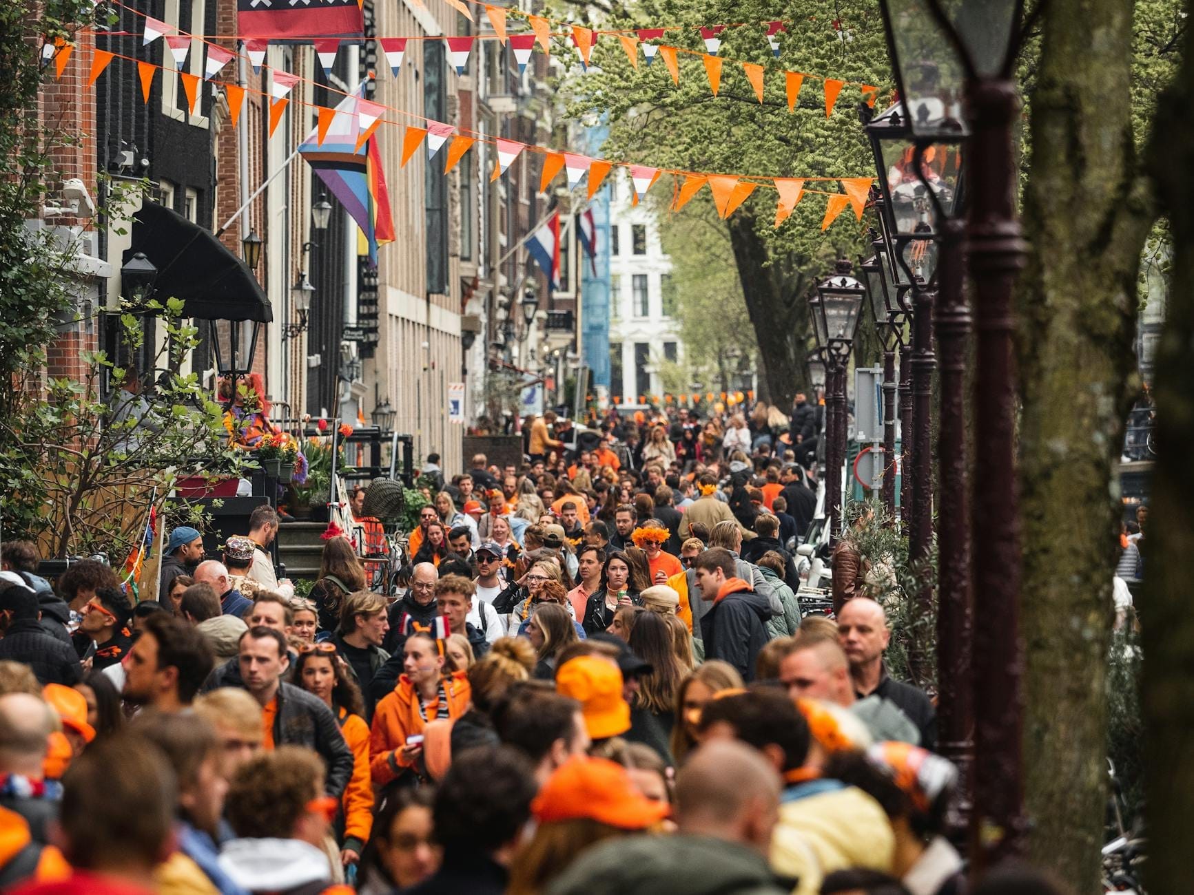 Calle concurrida de Amsterdam con gente vestida de naranja, banderines festivos en lo alto y un animado ambiente de fiesta callejera.