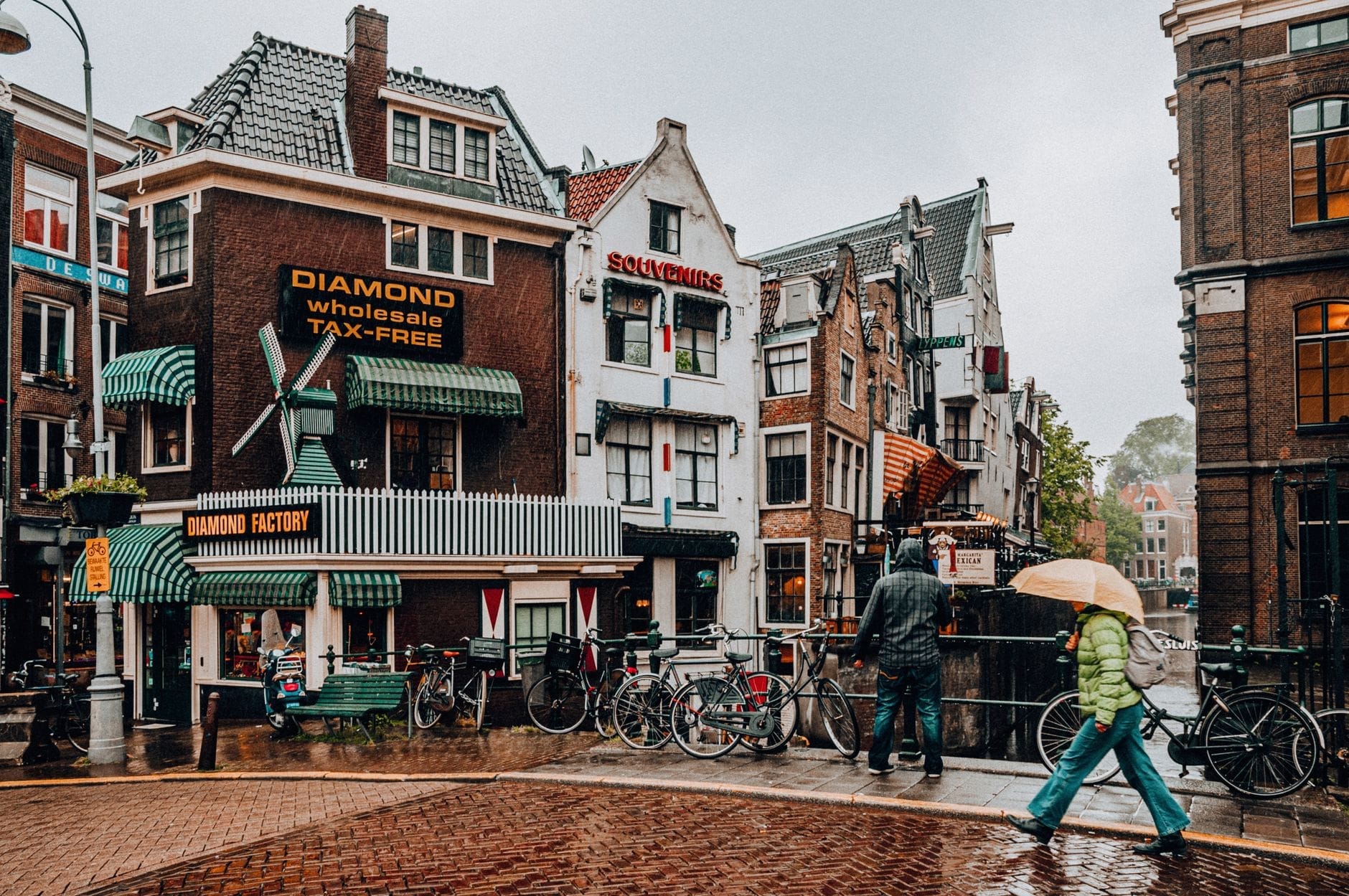 Escena de calle lluviosa en Ámsterdam con casas tradicionales junto al canal, personas caminando bajo paraguas y reflejos en el pavimento mojado.