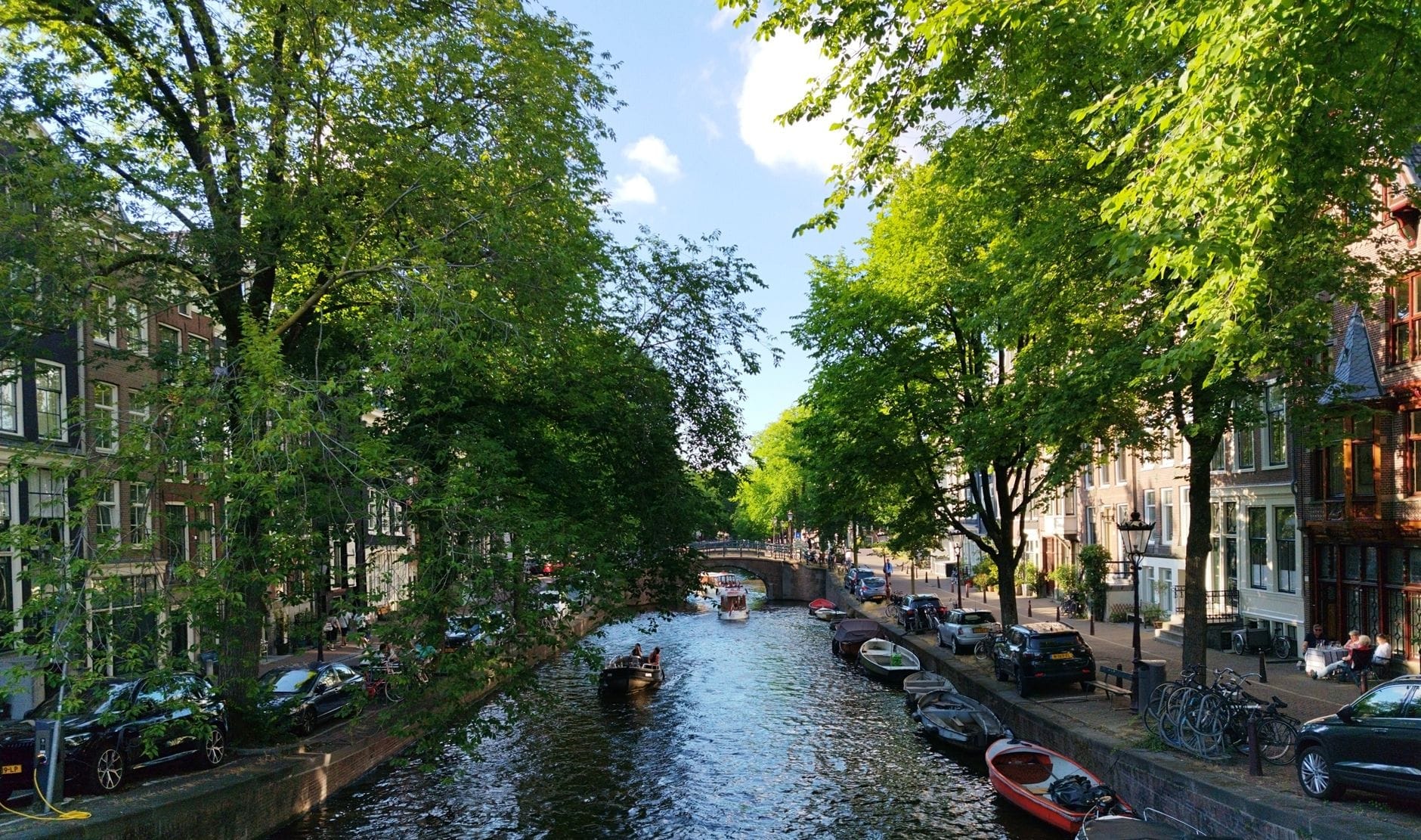Canal de Ámsterdam en un día soleado, bordeado de árboles verdes, barcas y casas típicas holandesas que transmiten el ambiente animado de la ciudad en su mejor tiempo.