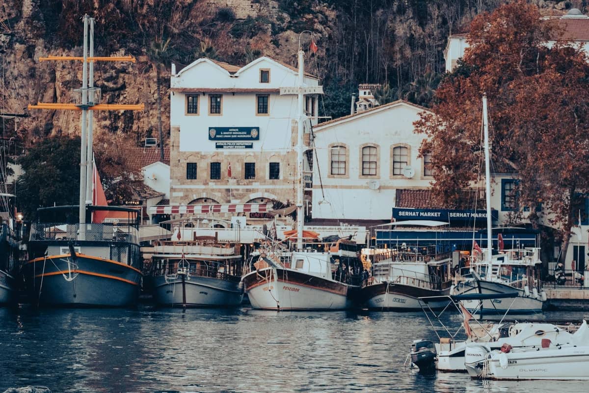Boats docked in Antalya's old harbor with autumn-colored trees and historic buildings in the background.