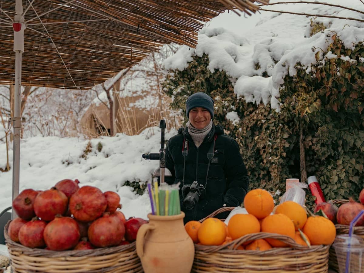 A smiling vendor stands behind baskets of oranges and pomegranates at an outdoor market, with snow-covered bushes and ground around, evoking winter freshness.