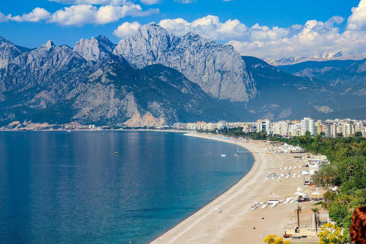 Wide photo of Antalya coastline with a sandy beach, clear blue sea, city buildings, and dramatic mountains under a partly cloudy sky.