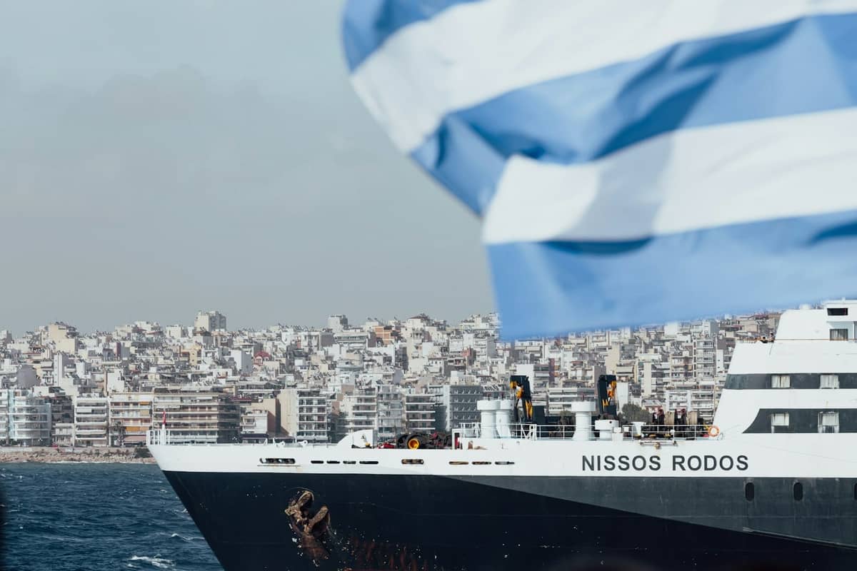 Zbliżenie dużego promu o nazwie Nissos Rodos w porcie Pireus w Atenach, z grecką flagą i budynkami miasta w tle.